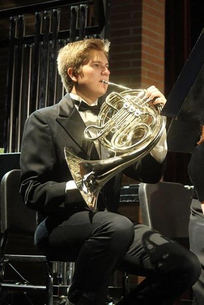 Dane Burton plays a French horn. He wears a black suit, and has light brown hair.